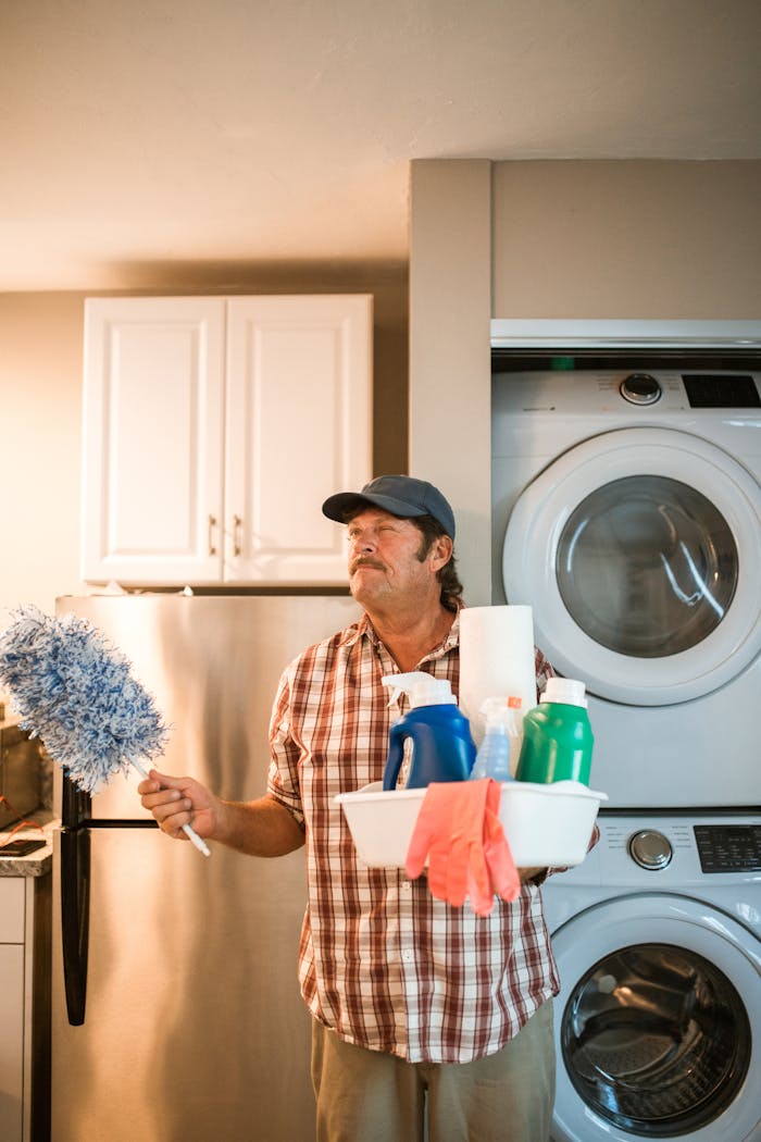 Adult man holding cleaning supplies next to washing machine in laundry room.
